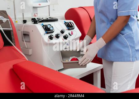 cropped view of multiracial nurse in blue uniform and latex gloves holding medical tray with rubber ball near transfusion machine in blood donation ce Stock Photo