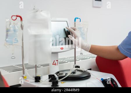 cropped view of multiracial nurse in latex glove operating transfusion machine with touchscreen near drip stands with infusion bags in blood donation Stock Photo