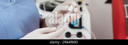 partial view of multiracial healthcare worker in blue uniform and latex gloves holding sterile test tube near blood transfusion machine on blurred bac Stock Photo