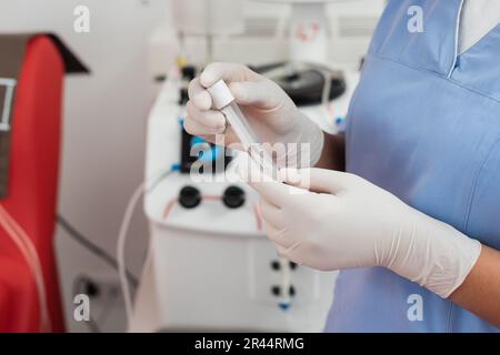 partial view of multiracial healthcare worker in blue uniform and latex gloves holding test tube near automated transfusion machine on blurred backgro Stock Photo