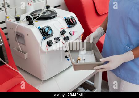 partial view of multiracial healthcare worker in uniform and latex gloves holding medical tray with test tubes near transfusion machine in blood donat Stock Photo