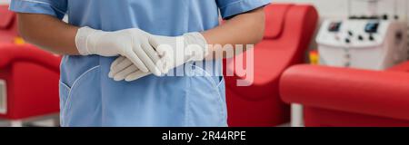 cropped view of multiracial healthcare worker in blue uniform and latex gloves standing in sterile environment of blood transfusion center, banner Stock Photo