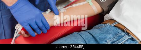 partial view of healthcare worker in latex gloves sticking band-aid on arm of multiracial woman sitting on medical chair with blood transfusion set, m Stock Photo