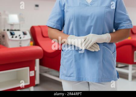 partial view of multiracial healthcare worker in blue uniform and latex gloves standing next to blurred medical chairs and transfusion machines in blo Stock Photo