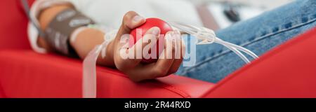 partial view of multiracial woman with transfusion set holding rubber ball while sitting on ergonomic medical chair during blood donation in laborator Stock Photo