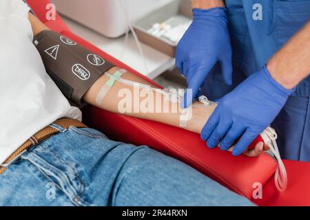 cropped view of doctor in latex gloves sticking band-aid on arm of multiracial woman with pressure cuff and transfusion set during blood donation in c Stock Photo