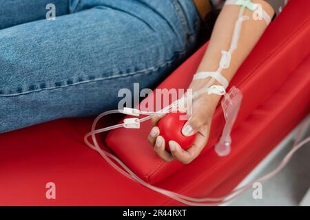 partial view of multiracial woman with a transfusion set holding rubber ball while sitting on ergonomic medical chair during blood donation in clinic, Stock Photo