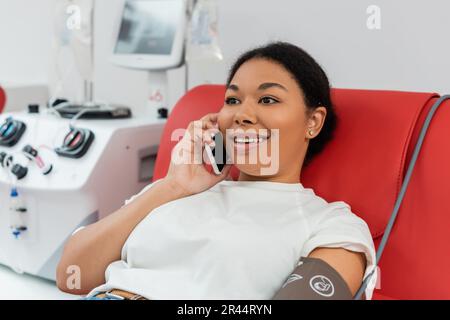 young and cheerful multiracial woman talking on smartphone while sitting on medical chair near blurred transfusion machine in blood donation center Stock Photo