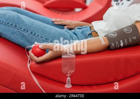 partial view of multiracial woman with transfusion set and medical rubber ball sitting on ergonomic medical chair during blood donation in laboratory Stock Photo