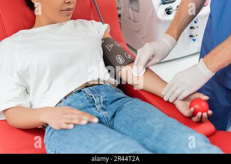 partial view of doctor in latex gloves holding alcohol pad and wiping arm of multiracial woman sitting with rubber ball on comfortable medical chair w Stock Photo