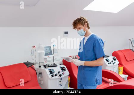 redhead doctor in blue uniform, medical mask and latex gloves looking at digital tablet near medical chairs and automated transfusion machines in bloo Stock Photo