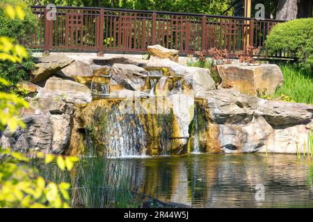 A small waterfall of a stream flowing into a garden pond, bordered by stone boulders along the coastline. Stock Photo