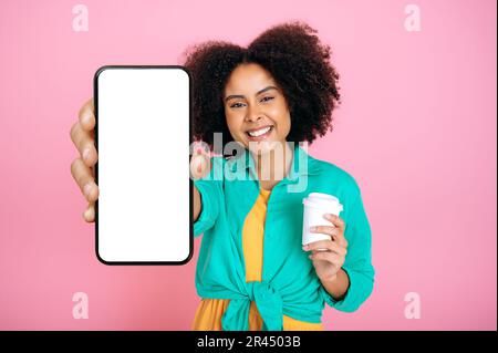 Lovely cheerful fashionable brazilian or hispanic woman, shows smartphone with white empty mock-up screen for advertisements or announcements, holds a coffee, smile at camera, isolated pink background Stock Photo