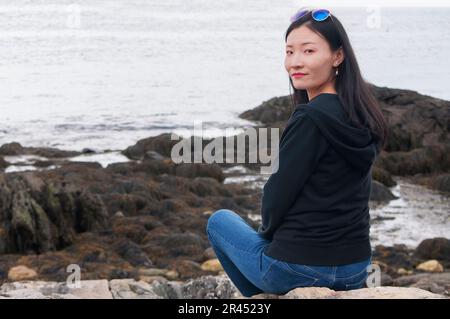 A chinese woman sitting on the rocky of shore of casco bay in Maine on an overcast day. Stock Photo