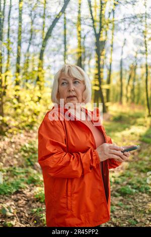 Fitness 50s 60s woman using smartphone, jogging in park and smiling, looking at her mobile phone app, checking sport application. Stock Photo