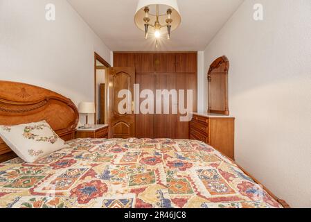 A bedroom with a double bed with a dresser with mirrored drawers and a built-in wardrobe with wooden doors Stock Photo