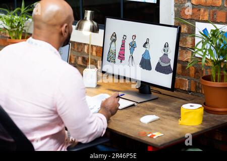 Biracial male fashion designer making fashion projects on computer in creative office, unaltered Stock Photo