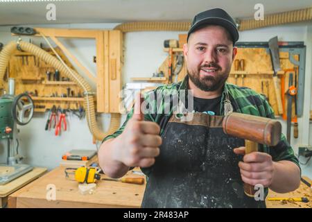 Smiling carpenter holding a hammer makes thumbs up sign in a woodworking studio. High quality photo Stock Photo