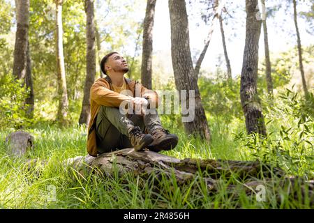Happy biracial man sitting on tree trunk and looking away in forest, unaltered Stock Photo