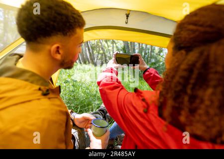 Happy biracial couple sitting in tent and using smartphone in forest, unaltered Stock Photo