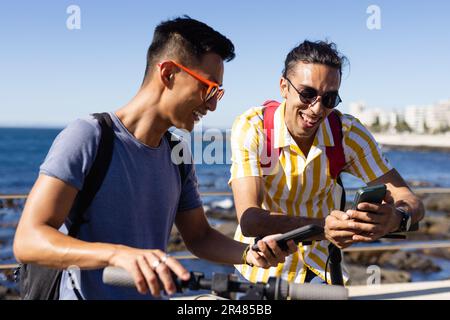 Happy biracial gay male couple using smartphone on promenade by the sea Stock Photo