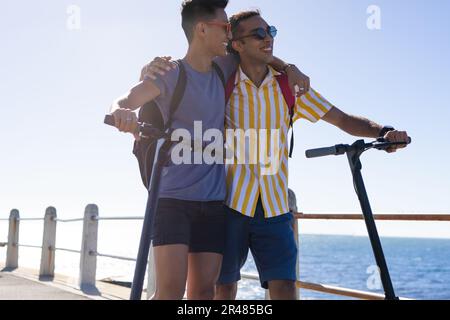 Happy biracial gay male couple walking with scooters embracing on promenade by the sea Stock Photo