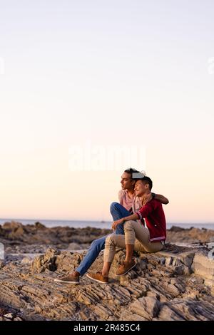 Happy biracial gay male couple sitting on rocks and embracing on beach at sundown, with copy space Stock Photo