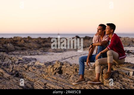 Happy biracial gay male couple sitting on rocks and embracing on beach at sundown, with copy space Stock Photo