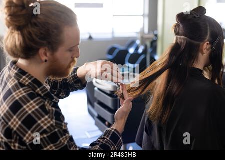 Focused caucasian male hairdresser with hair bun cutting female customer's long hair at salon Stock Photo