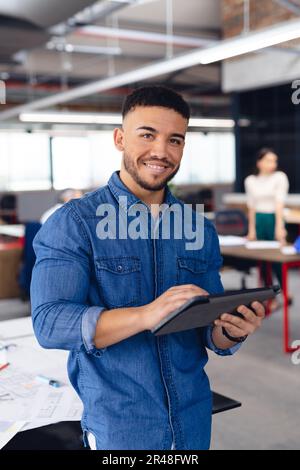 Portrait of happy biracial businessman using tablet and smiling at office, unaltered Stock Photo