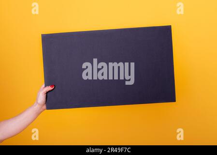 Female hand holding a blank black sheet poster for copy-space and mockup on a yellow background. Stock Photo