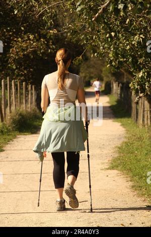 A woman in activewear and walking sticks is walking down a path of dirt. Stock Photo