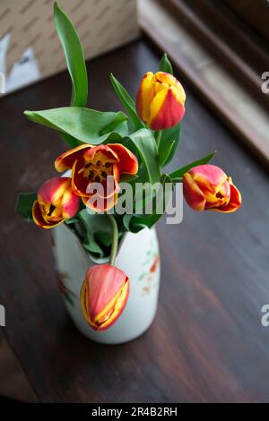 Top view of a white porcelain jug containing a bouquet of red and yellow tulips on a wooden table. Vertical image with selective focus and shallow dep Stock Photo