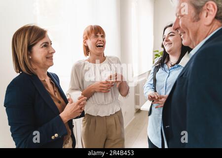 Colleagues of mixed ages and genders taking a break in office, standing near a window, laughing and chatting. Focus is on young redhead woman laughing Stock Photo