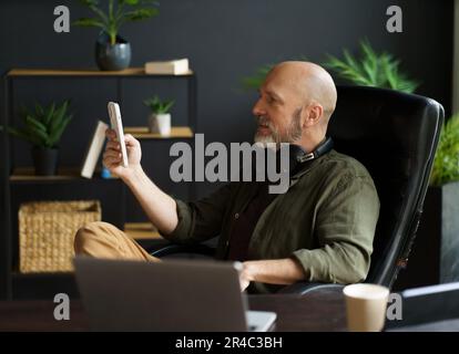 Smart and tech-savvy mid-aged man sits in chair at desk, engrossed in screen of phone. With bald head, silver beard, and air of handsomeness, multitasks, balancing phone, laptop, and cup of coffee. High quality photo Stock Photo