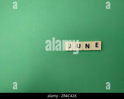 A close-up of a set of vintage-style Scrabble tiles forming the word 'June' against a green wall Stock Photo