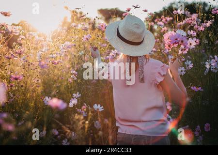 A female with a fashionable hat stands in a lush meadow filled with vibrant flowers Stock Photo