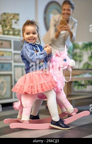 Lovely toddler girl riding a pink unicorn rocking horse in a shop, feeling joyful, mother taking a picture in the background. Shopping, family, lifest Stock Photo