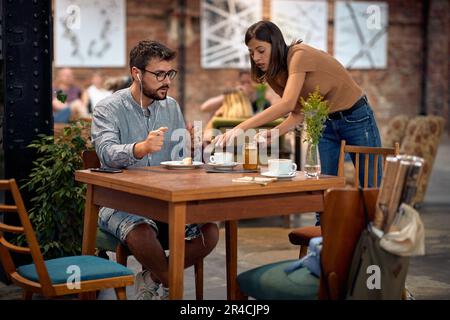 Young students in cafeteria, enjoying coffee. Woman standing and managing things on table, cleaning with paper towel. Youth, students, cafe, lifestyle Stock Photo
