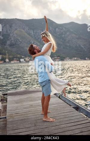 Young bride, fashionable blonde in beautiful dress with groom on dock by sea. Man holding her, she feels love, joy and freedom. Wedding, honeymoon, lo Stock Photo