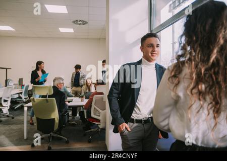 Two young business colleagues talking and smiling while standing next to the windows of the co working space Stock Photo