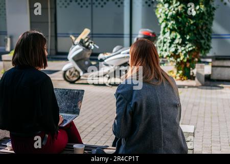 Back view shot of two businesswomen sitting on a bench in the city and having a conversation. Brunette girl holding a lap top on her lap Stock Photo