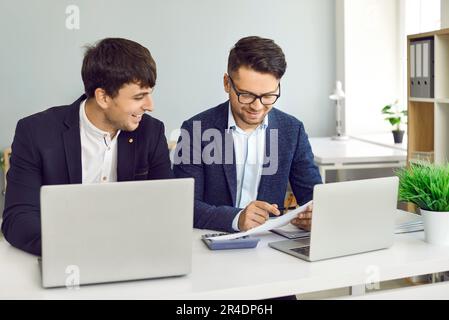 Smiling businesspeople using laptop, talking, communication, planning startup, working in office. Two business colleagues in meeting Stock Photo