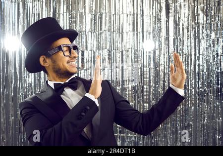 Smiling young latin american in black tuxedo holding empty space on shiny silver background. Stock Photo