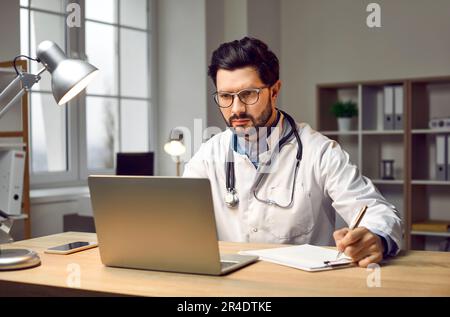 Male doctor sitting in office, working on laptop computer, doing research and taking notes Stock Photo