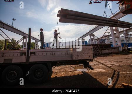 Podolsk, Moscow province - August 02, 2021: Silhouettes of workers loading pipes with overhead crane. Pipes warehouse. Stock Photo