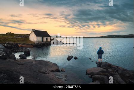 Magical nature scenery with man standing on the rock by the lake and watching sunrise behind fisherman's hut at Screebe, connemara national Park in County Galway, Ireland Stock Photo