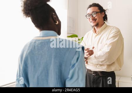 A young Chinese man, holding a disposable cup, chatting with his African colleague during a break in a modern, bright office. Scene is lit by a window Stock Photo