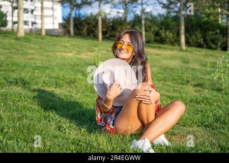 Joyful woman with sunglasses looking at camera and smiling while sitting outdoors on a green grass in a park. Stock Photo