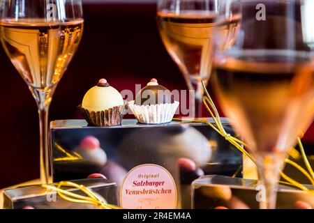 Two original Salzburg Venusbrüstchen (Nipples of Venus) rest on a gift box in Salzburg, Austria, surrounded by glasses of sparkling wine. The pralines are made of chestnut nougat cream and a piece of marinated Amarena cherry, coated in white or dark chocolate. Recommended pairing: the white ones with Prosecco, the dark ones with coffee. Stock Photo